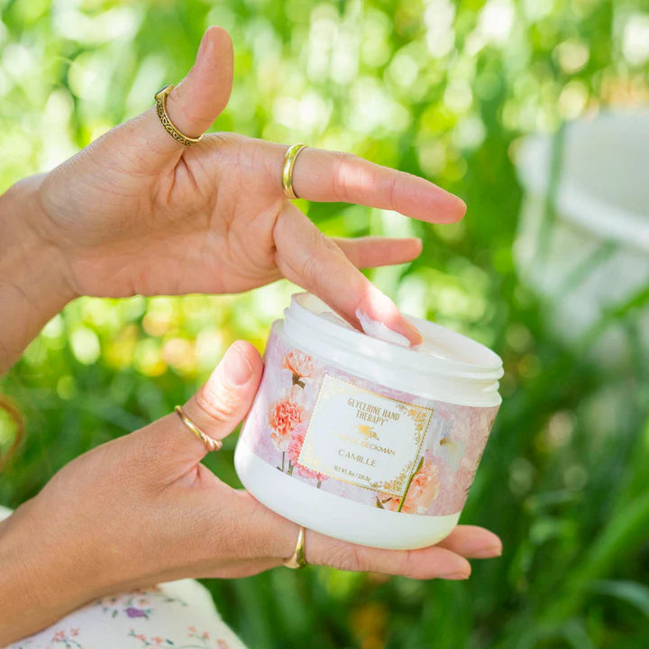 A woman holds Camille Beckman's Glycerine Hand Therapy in front of outdoor greenery. 